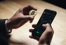 Hand holding a physical Bitcoin coin next to a phone showing a crypto trading app