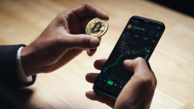 Hand holding a physical Bitcoin coin next to a phone showing a crypto trading app